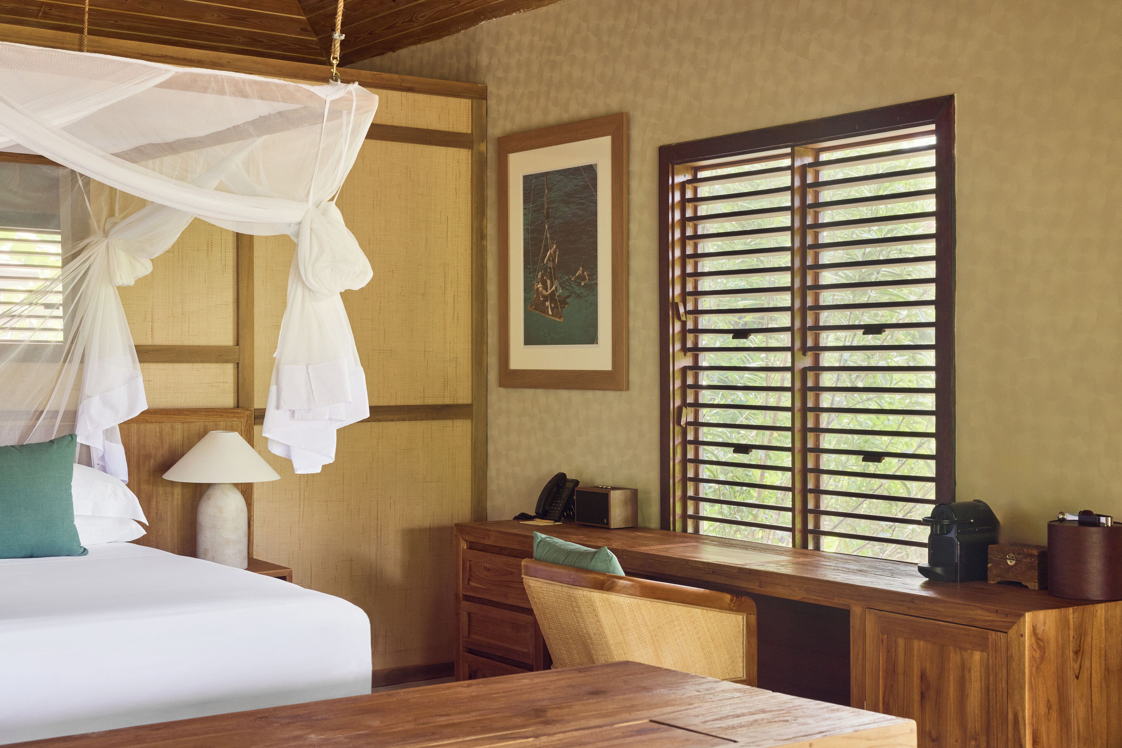 A hotel room with a bed, a desk, a chair, a lamp, a telephone, and a window with blinds.