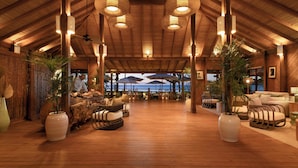 A spacious indoor area with wooden flooring, large potted plants, and a view of the ocean through large windows.