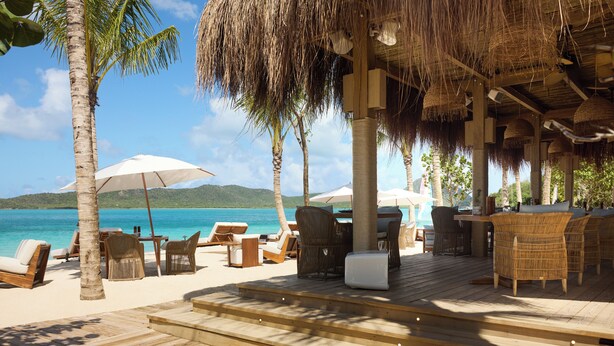 A beachside lounge with wicker furniture, umbrellas, and a thatched roof structure.