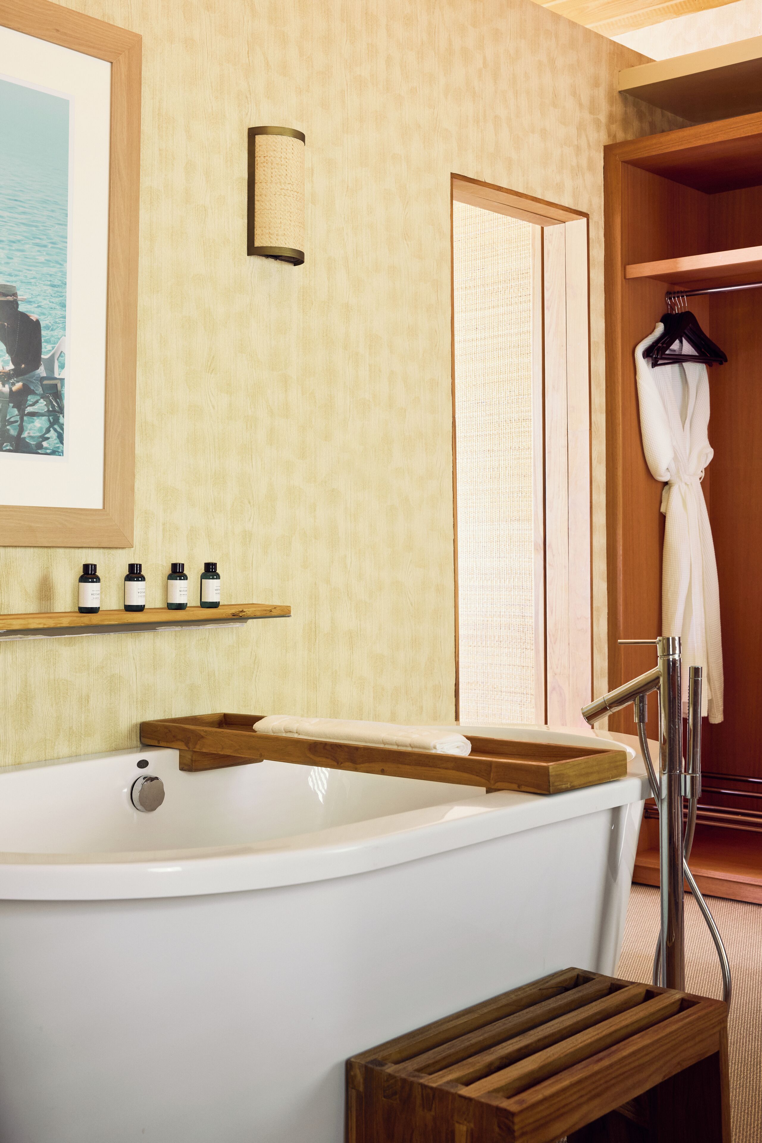 A bathroom with a freestanding bathtub, wooden shelves with bottles, a towel rack with a robe, and a framed picture on the wall.