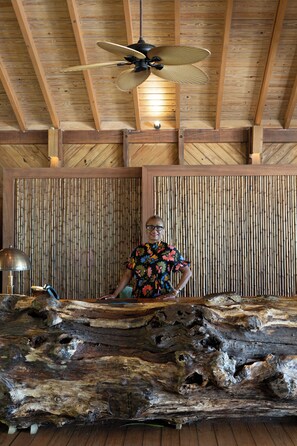 A room with a wooden ceiling, a large fan, bamboo wall panels, and a rustic wooden table.