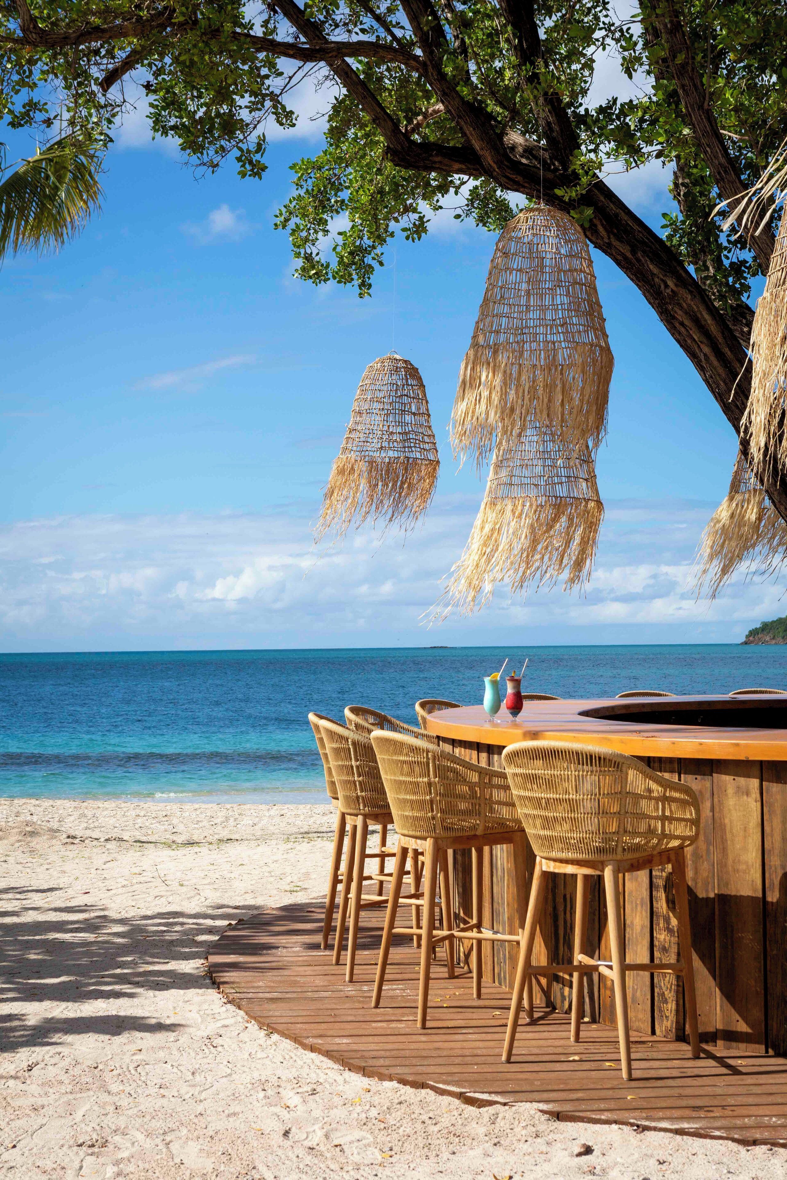 A wooden deck with wicker chairs and a table, overlooking a beach with clear blue water.
