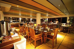 Uma ampla sala de jantar com várias mesas e cadeiras, um serviço de buffet e uma área de cozinha ao fundo.