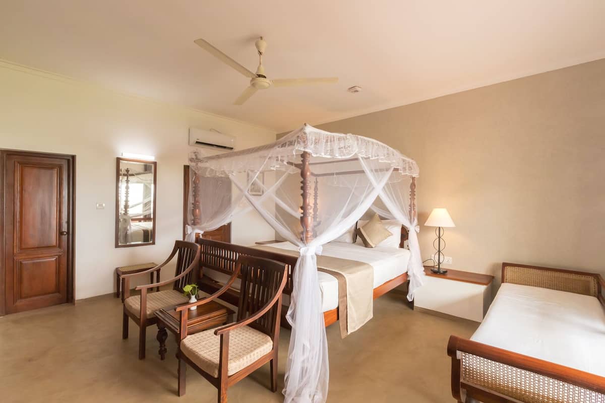 A bedroom with a four-poster bed, a ceiling fan, a bedside table with a lamp, and two wooden chairs.
