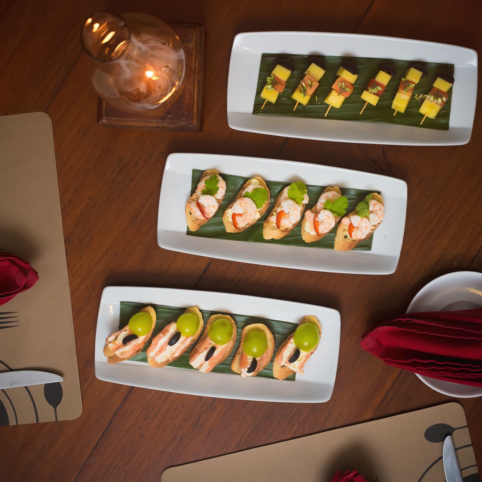 Une table avec trois assiettes rectangulaires servant de présentoir pour des amuse-gueules, une bougie et une serviette.