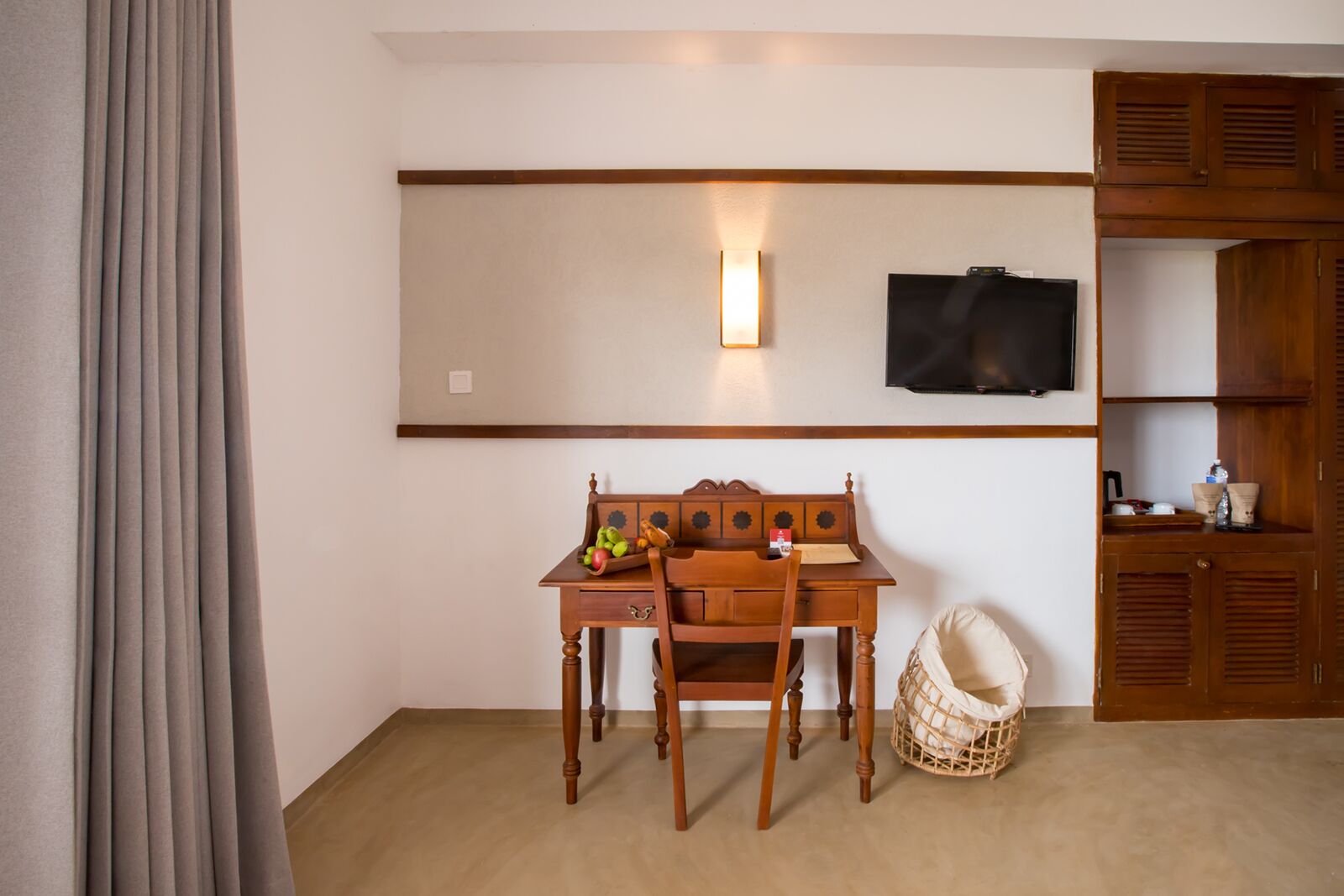 A room with a wooden desk, a chair, a television mounted on the wall, and a wooden wardrobe.