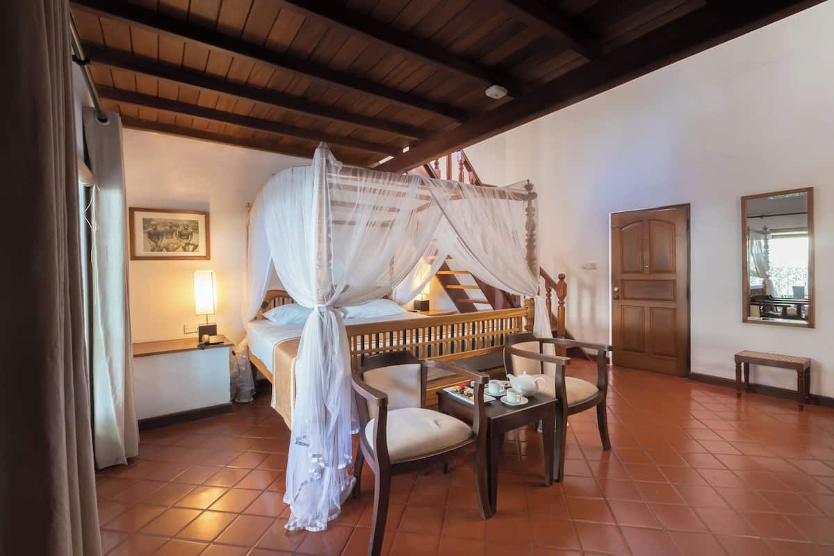A room with a bed under a canopy, a wooden chair, a small table with a teapot, and a wooden door.