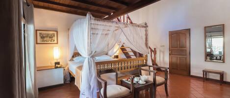 A room with a bed under a canopy, a wooden chair, a small table with a teapot, and a wooden door.