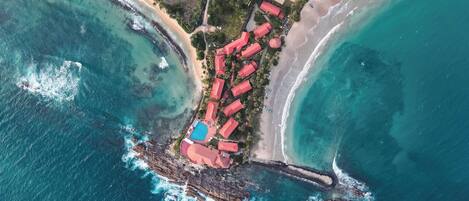Vista aérea de un complejo turístico costero con varias edificaciones de techo rojo, una piscina y una playa.