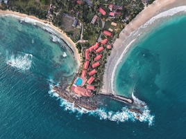 Vista aérea de um resort costeiro com vários edifícios de telhado vermelho, uma piscina e uma praia.