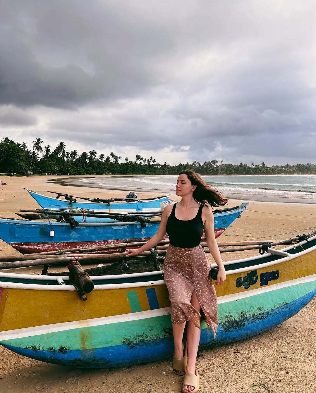 A woman standing next to a colorful boat on a beach with palm trees in the background.