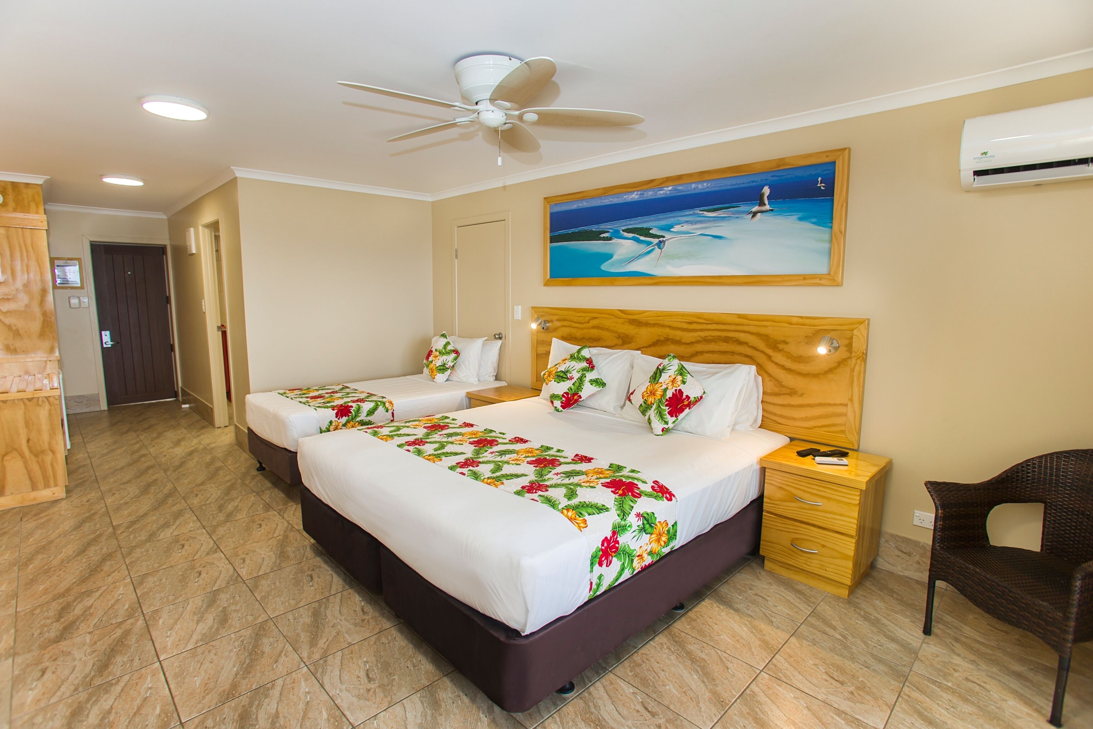 Ein Hotelzimmer mit zwei Betten, einem Deckenventilator, einer Klimaanlage, einem hölzernen Kopfteil, einem Nachttisch, einem Sessel und einem Gemälde mit Strandmotiv.