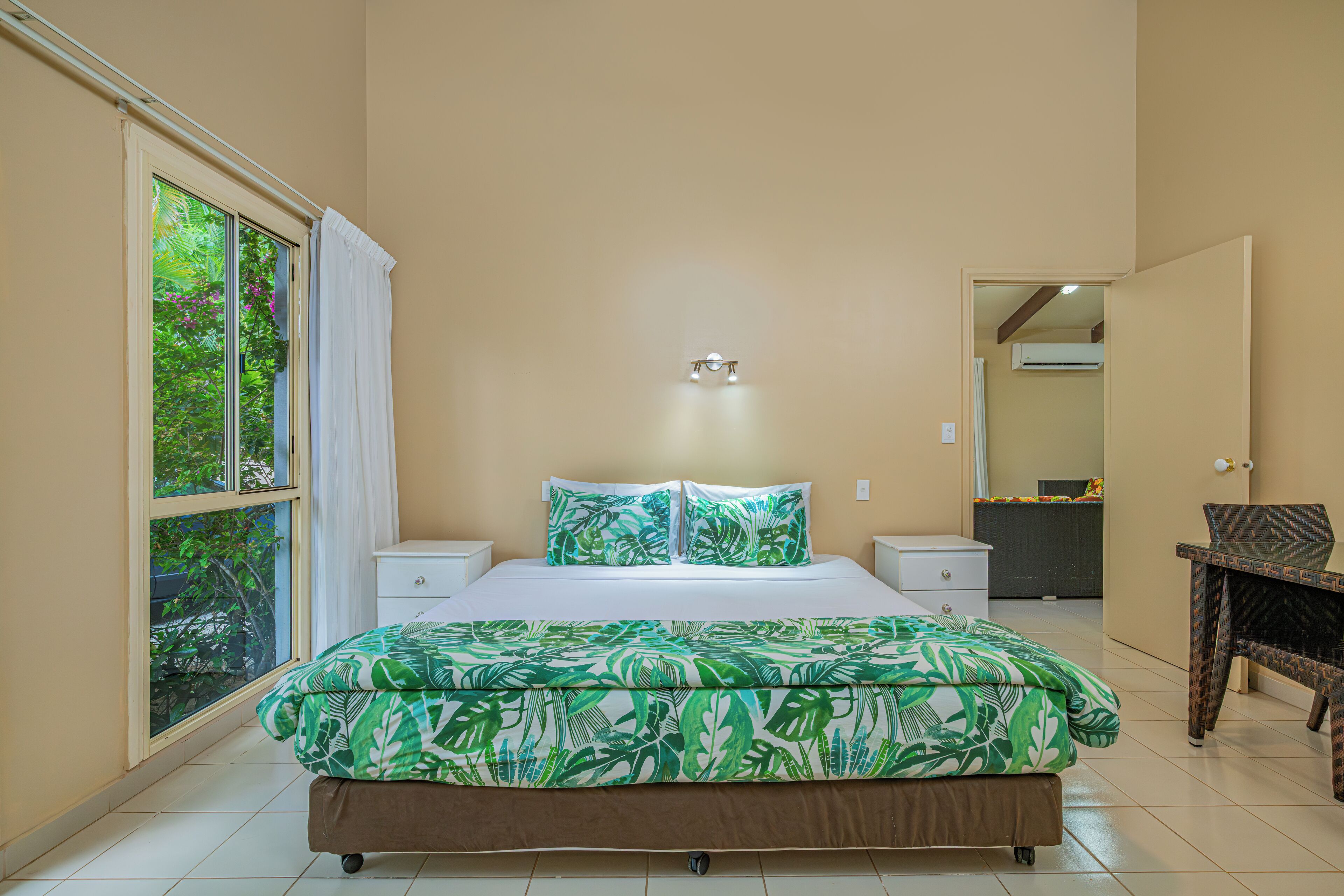 Un dormitorio con cama, mesitas de noche blancas, un escritorio y una ventana con cortinas.