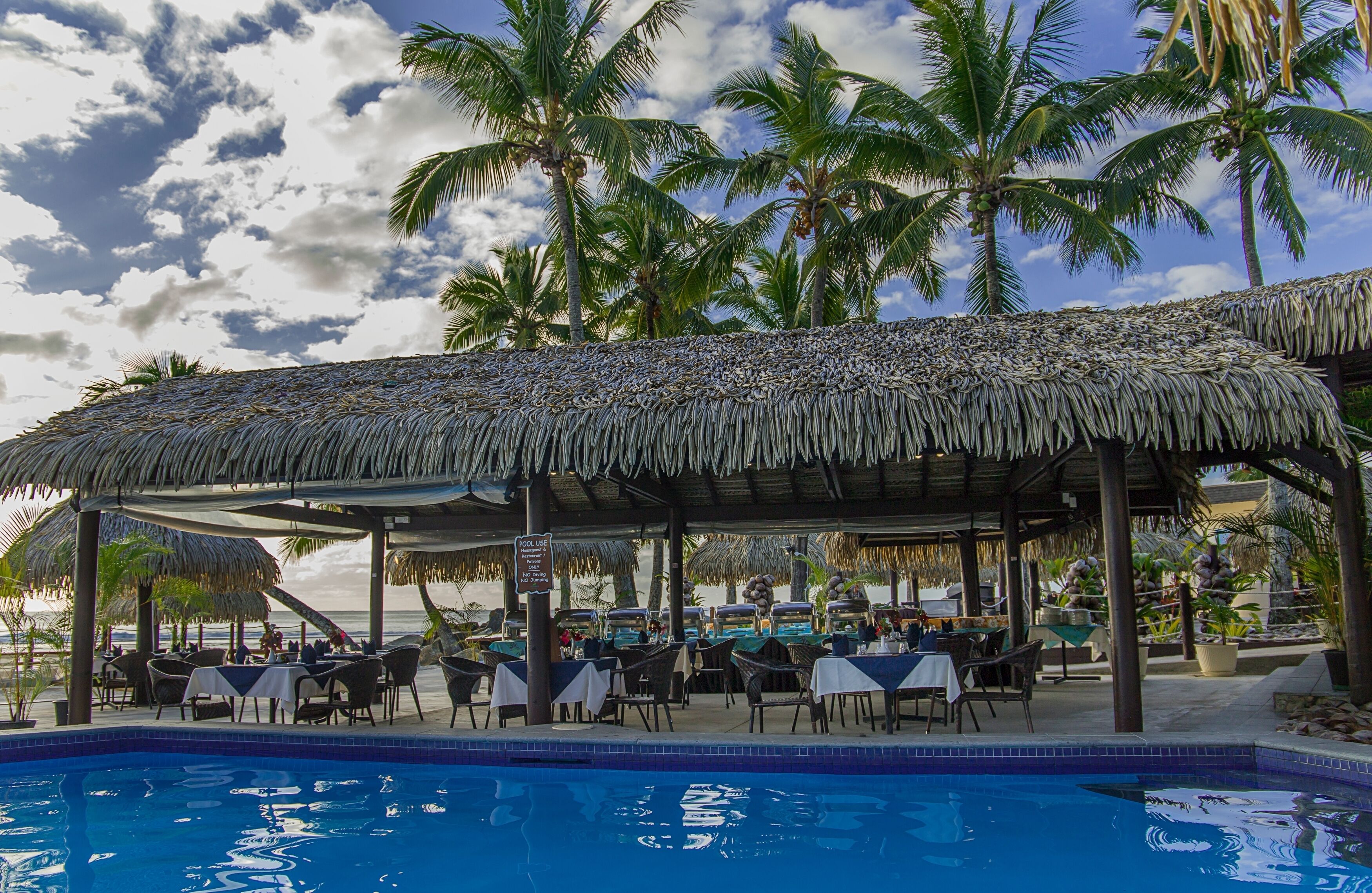 A poolside restaurant with thatched roofs, tables, and chairs, overlooking a pool and palm trees.