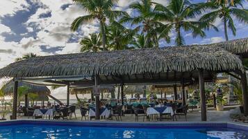 A poolside restaurant with thatched roofs, tables, and chairs, overlooking a pool and palm trees.