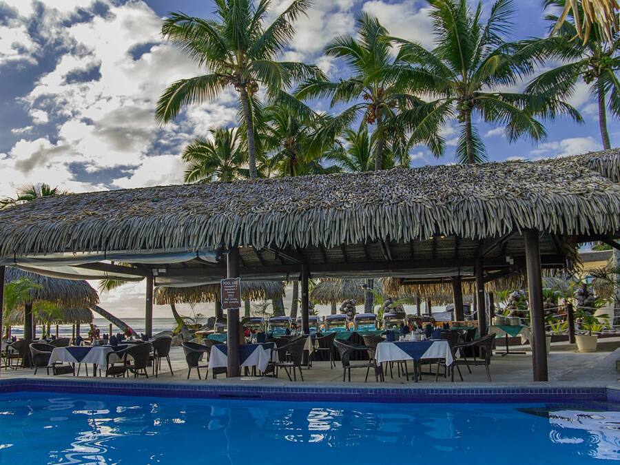 A poolside restaurant with thatched roofs, tables, and chairs, overlooking a pool and palm trees.
