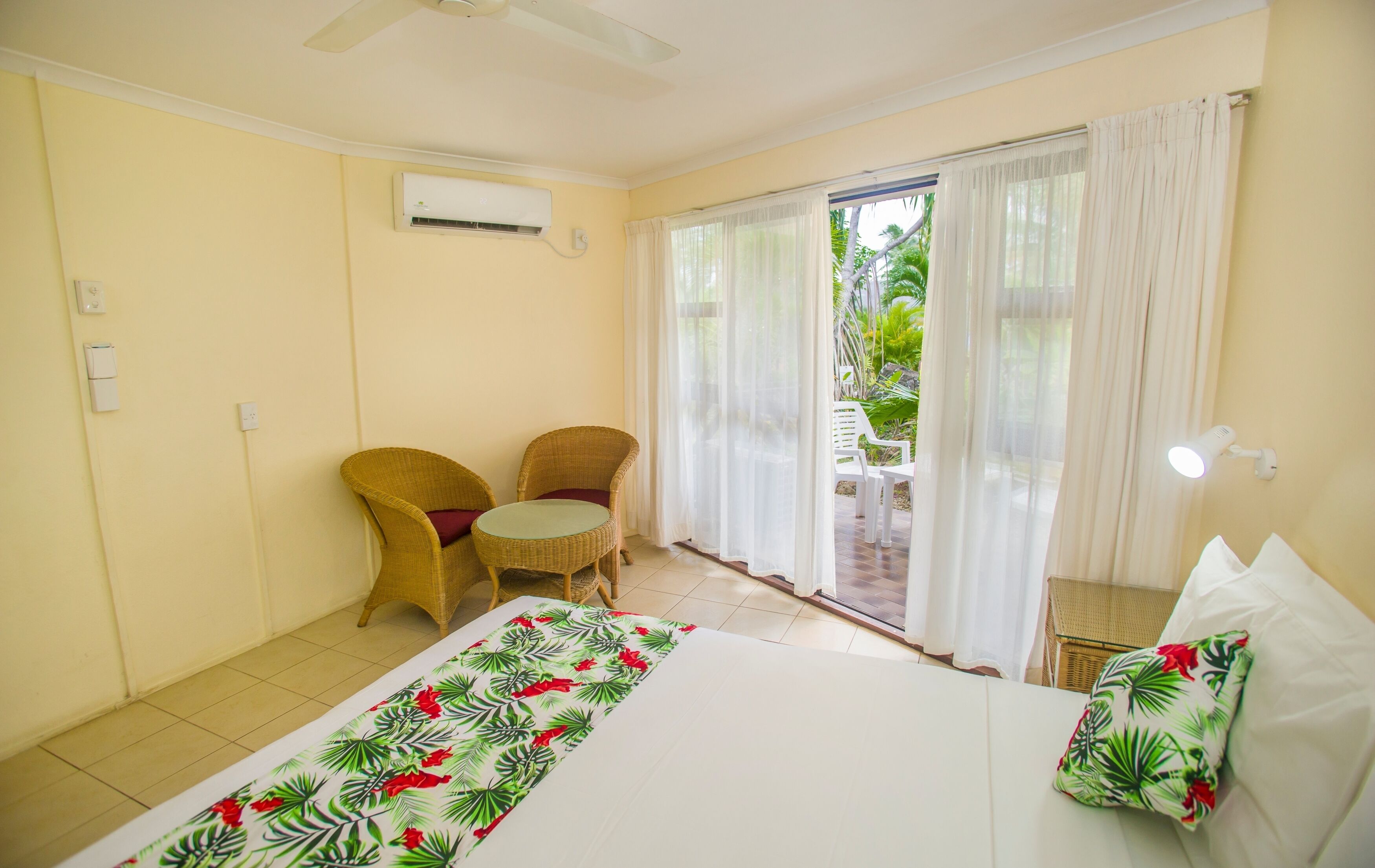 A hotel room with a bed, two wicker chairs, a small table, and a view of the outdoors.