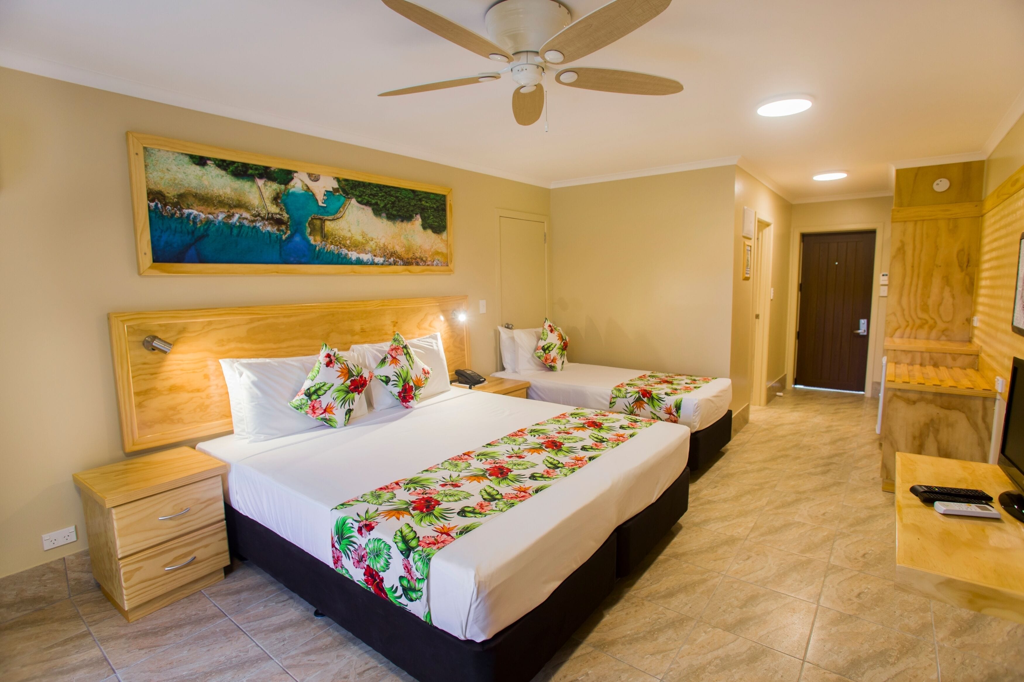 A hotel room with two beds, a wooden headboard, a painting on the wall, a ceiling fan, and a desk with a TV and remote controls.
