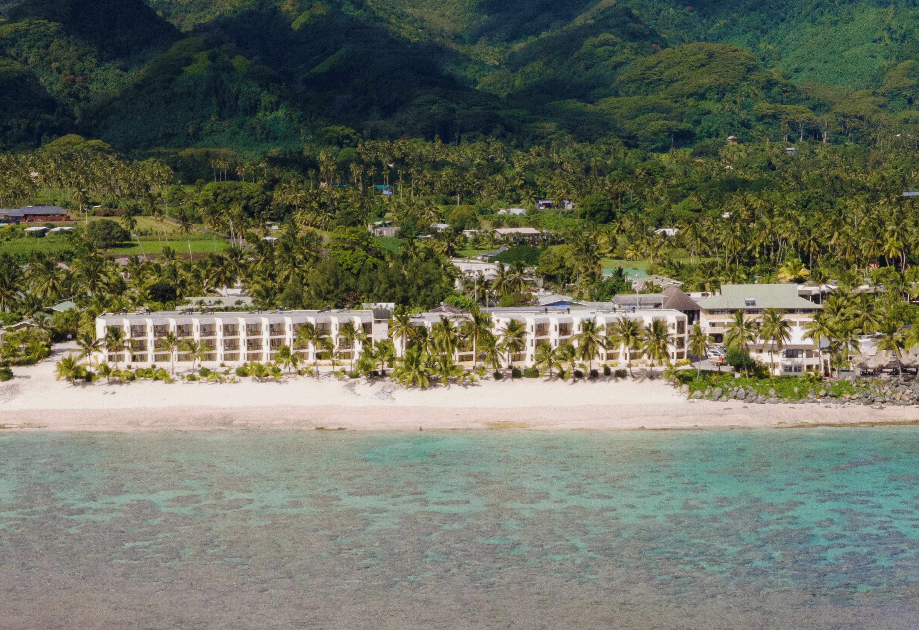 An aerial view of a resort with a beachfront, palm trees, and lush greenery in the background.