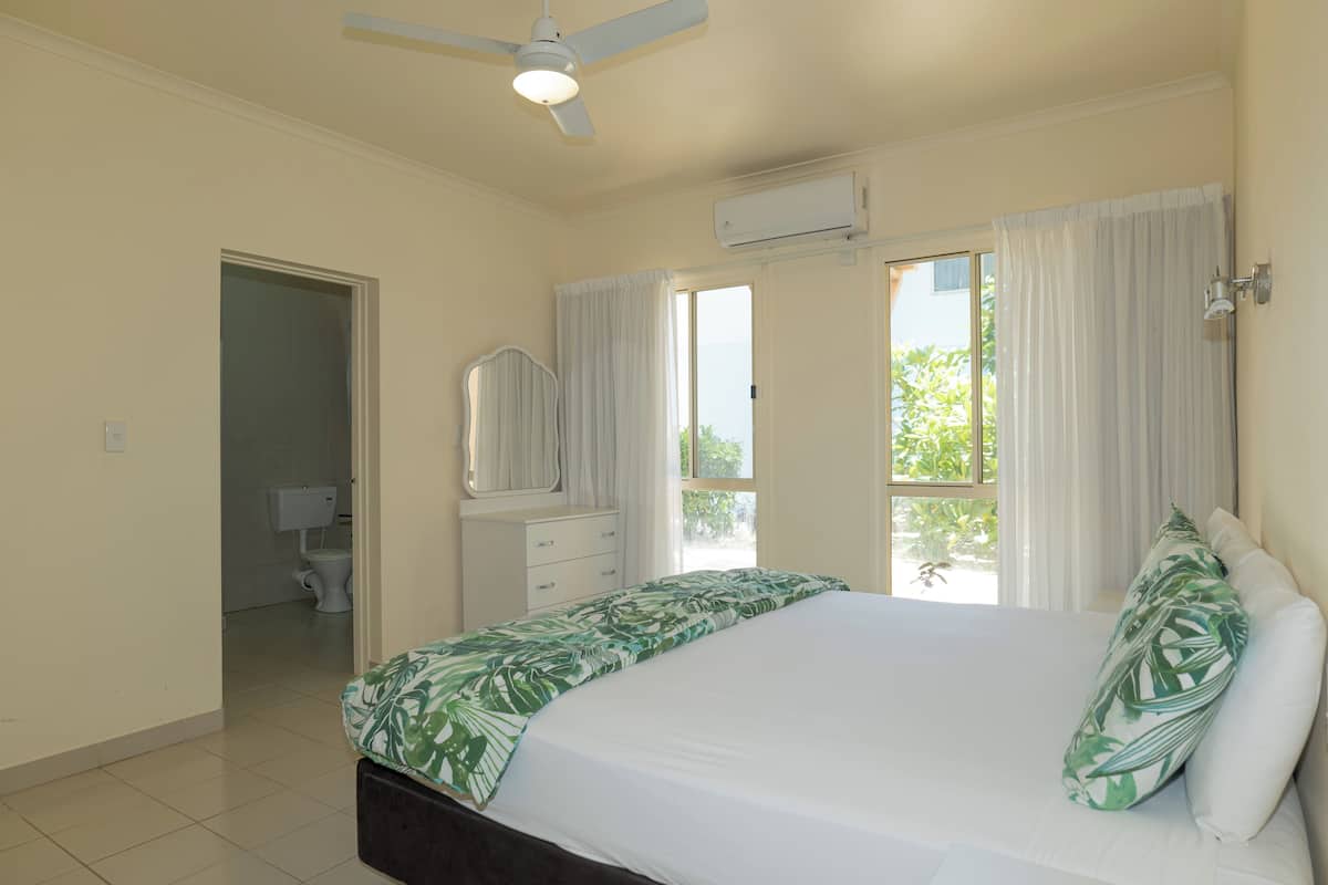 Une chambre à coucher avec un lit, un ventilateur de plafond, un appareil de climatisation, une fenêtre avec des rideaux et une salle de bain visible à travers une porte ouverte.