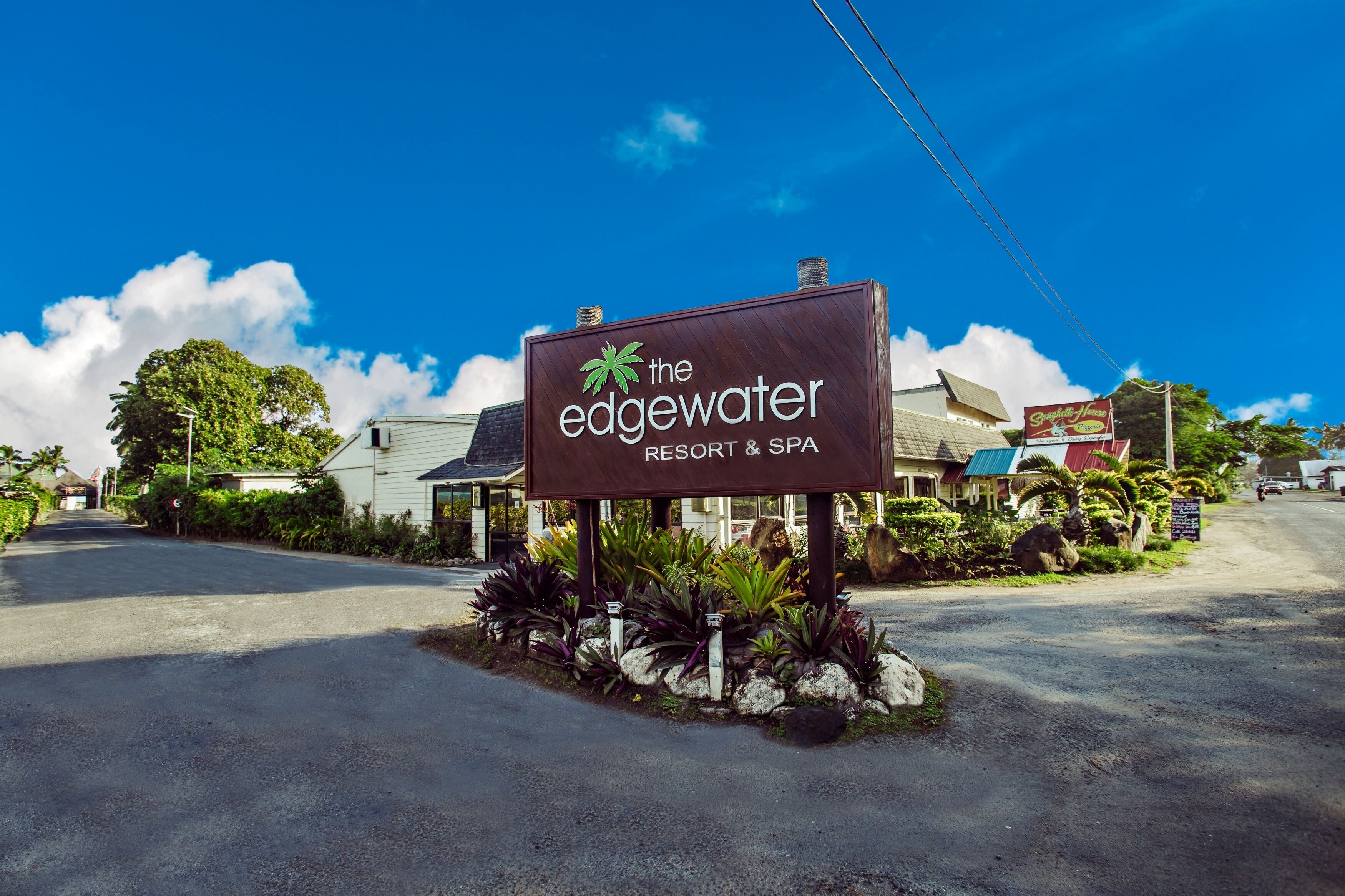 Une enseigne pour le complexe hôtelier et spa The Edgewater, arborant un thème tropical, entourée d’une végétation luxuriante et sous un ciel bleu azur.