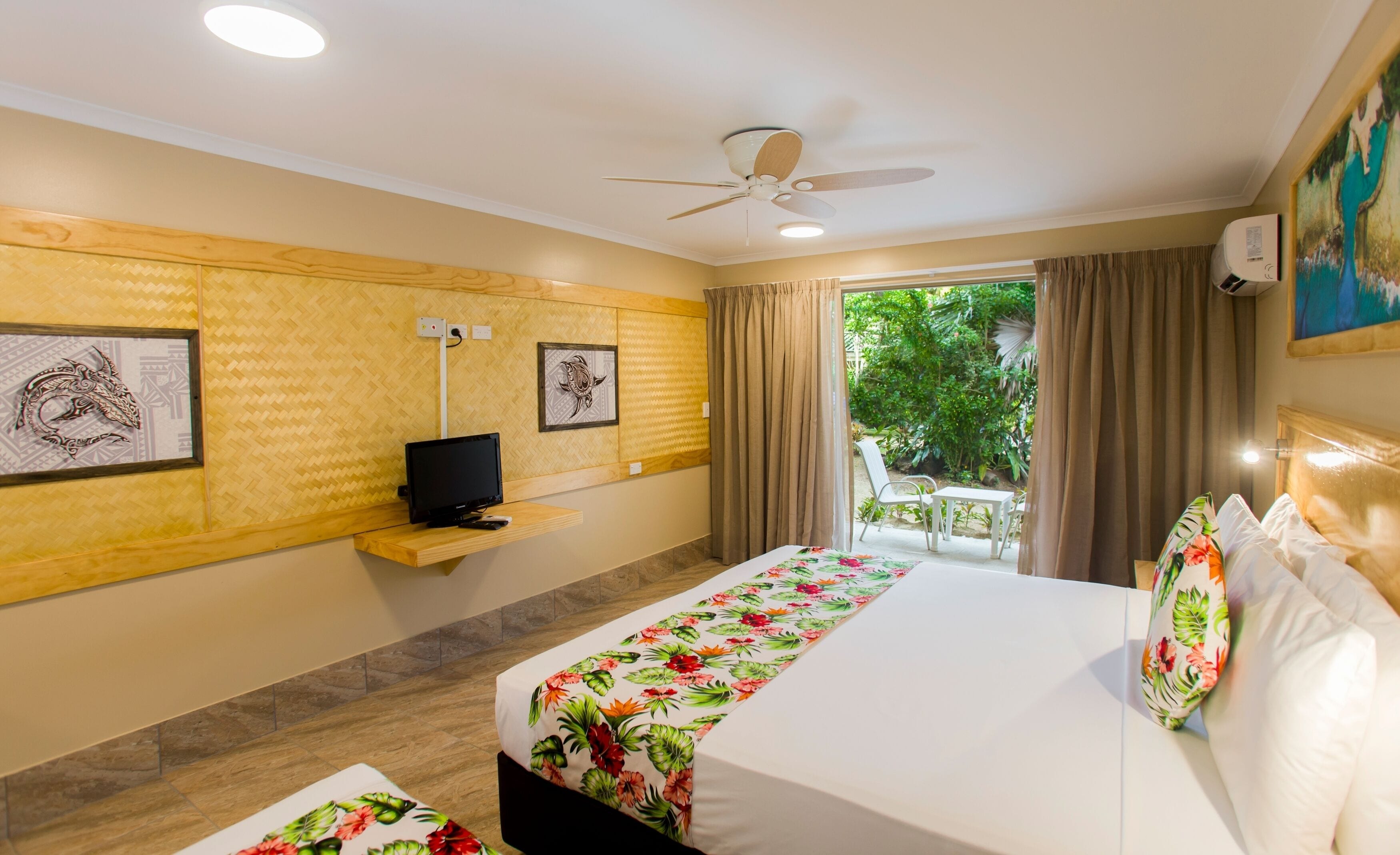 A hotel room with a bed, a TV, a ceiling fan, and a window with curtains.