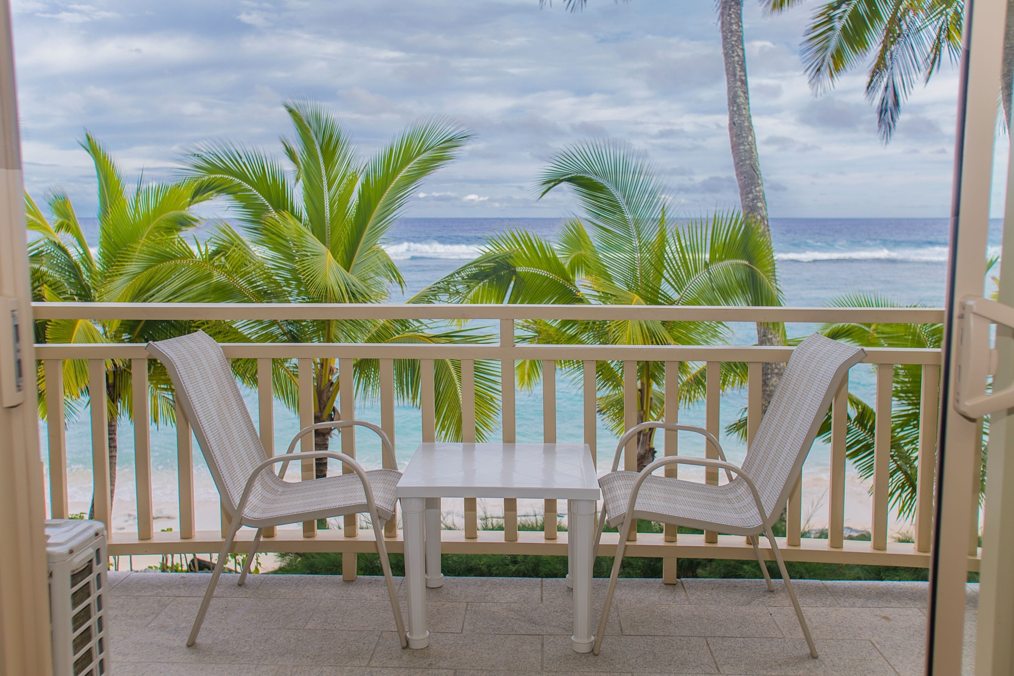 Een balkon met een tafel en twee stoelen, uitkijkend op een strand met palmbomen en de oceaan.