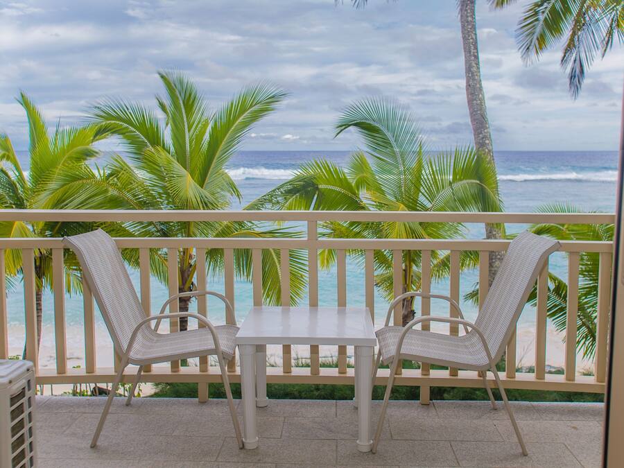 Een balkon met een tafel en twee stoelen, uitkijkend op een strand met palmbomen en de oceaan.