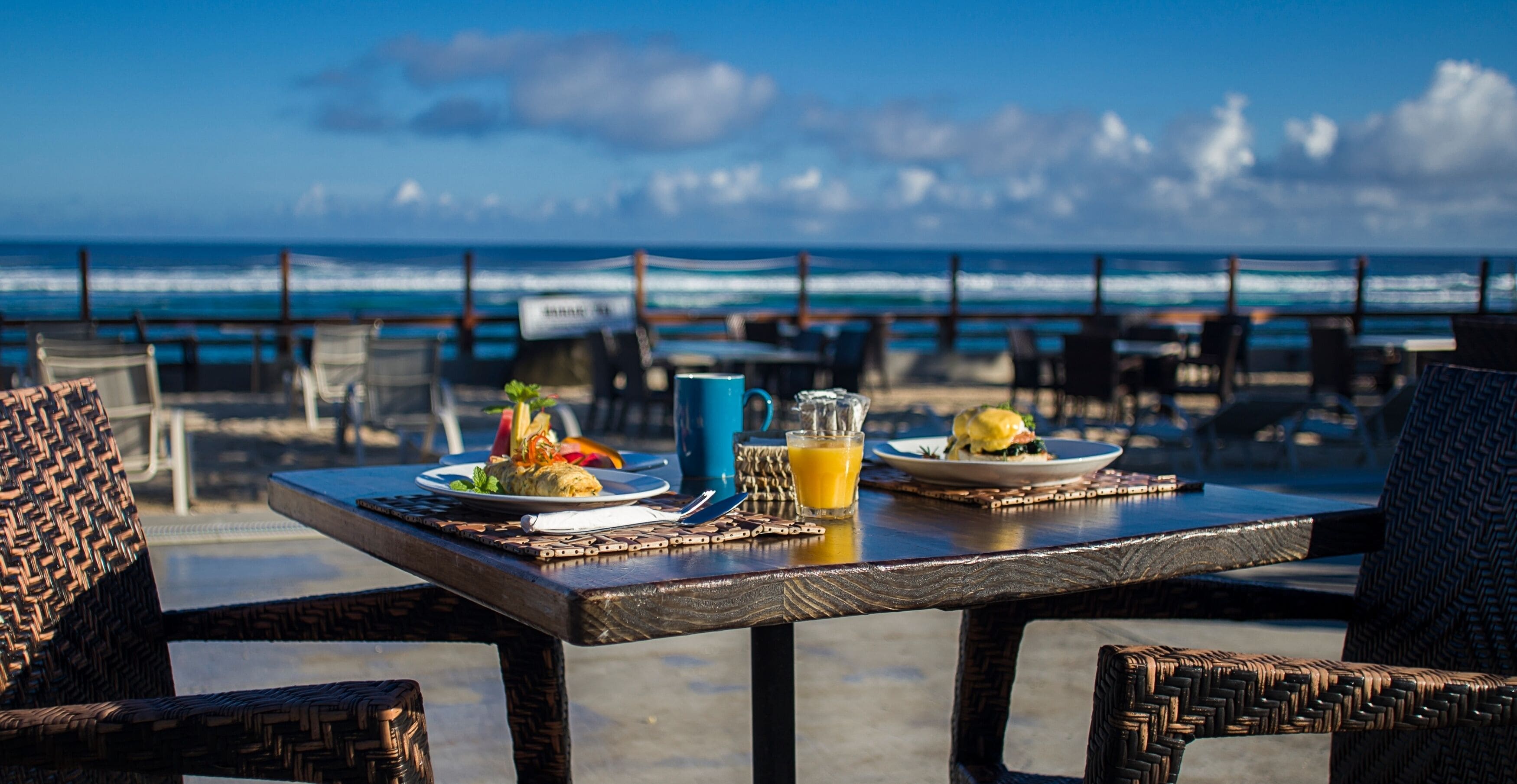 Mesa com itens de café da manhã e um copo de suco de laranja, com vista para o oceano.