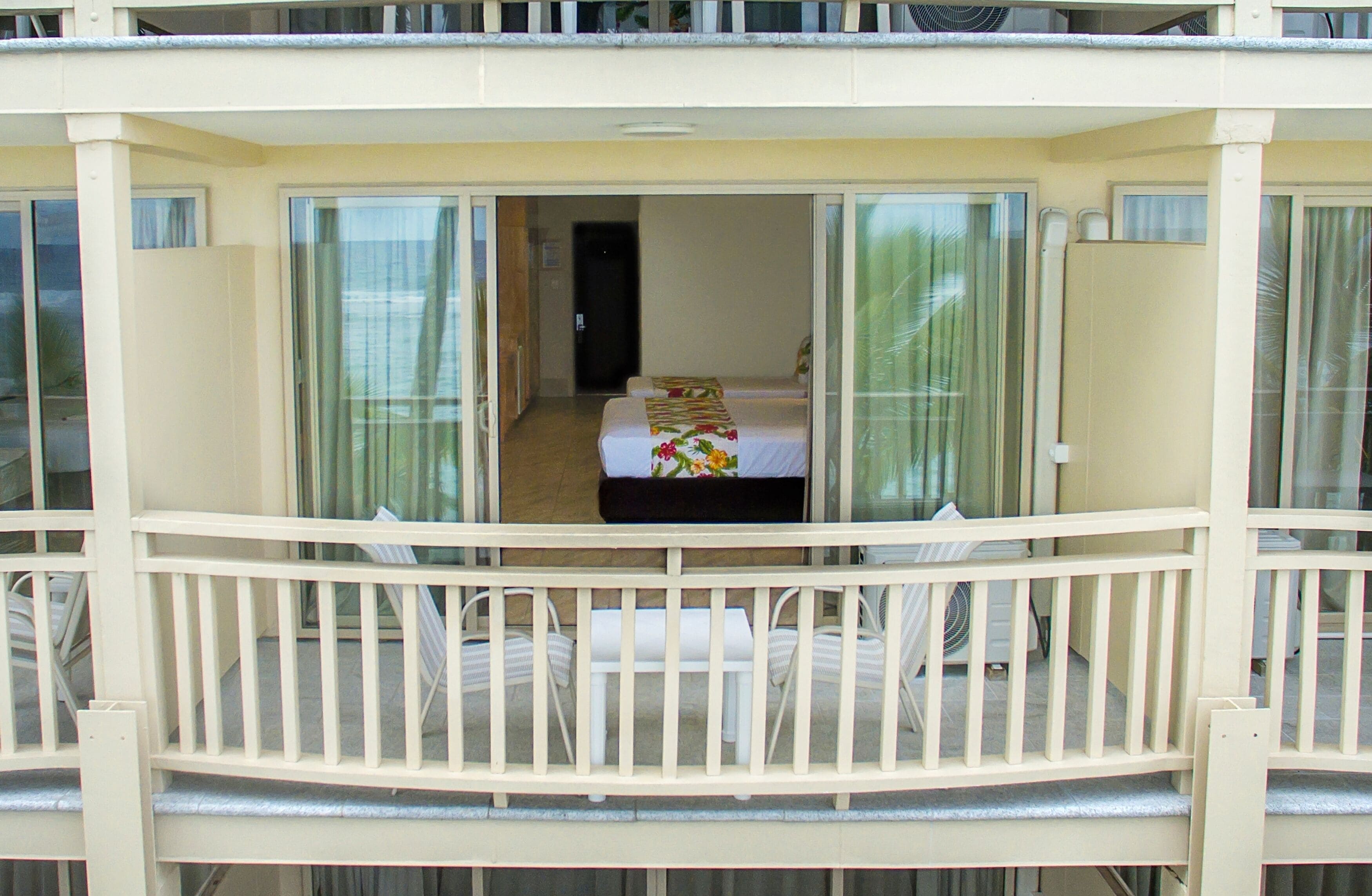 A balcony with a bed and a small table, a white railing, and a view of the interior.