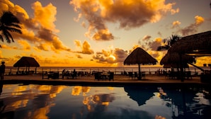A beachside pool area with thatched umbrellas, lounge chairs, and a view of the sunset over the ocean. - The Edgewater Resort & Spa (Rarotonga)