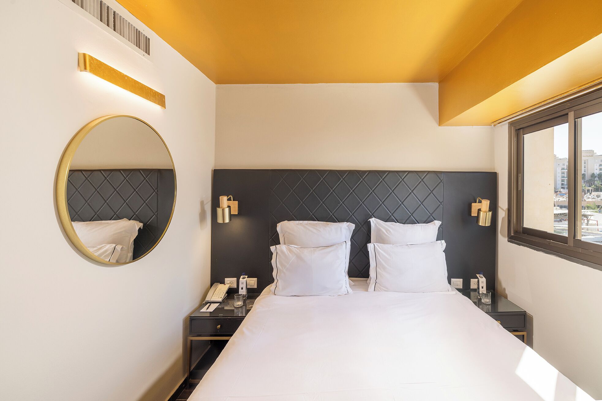 A hotel room with a large bed, a round mirror, bedside tables with lamps, and a window with a city view.