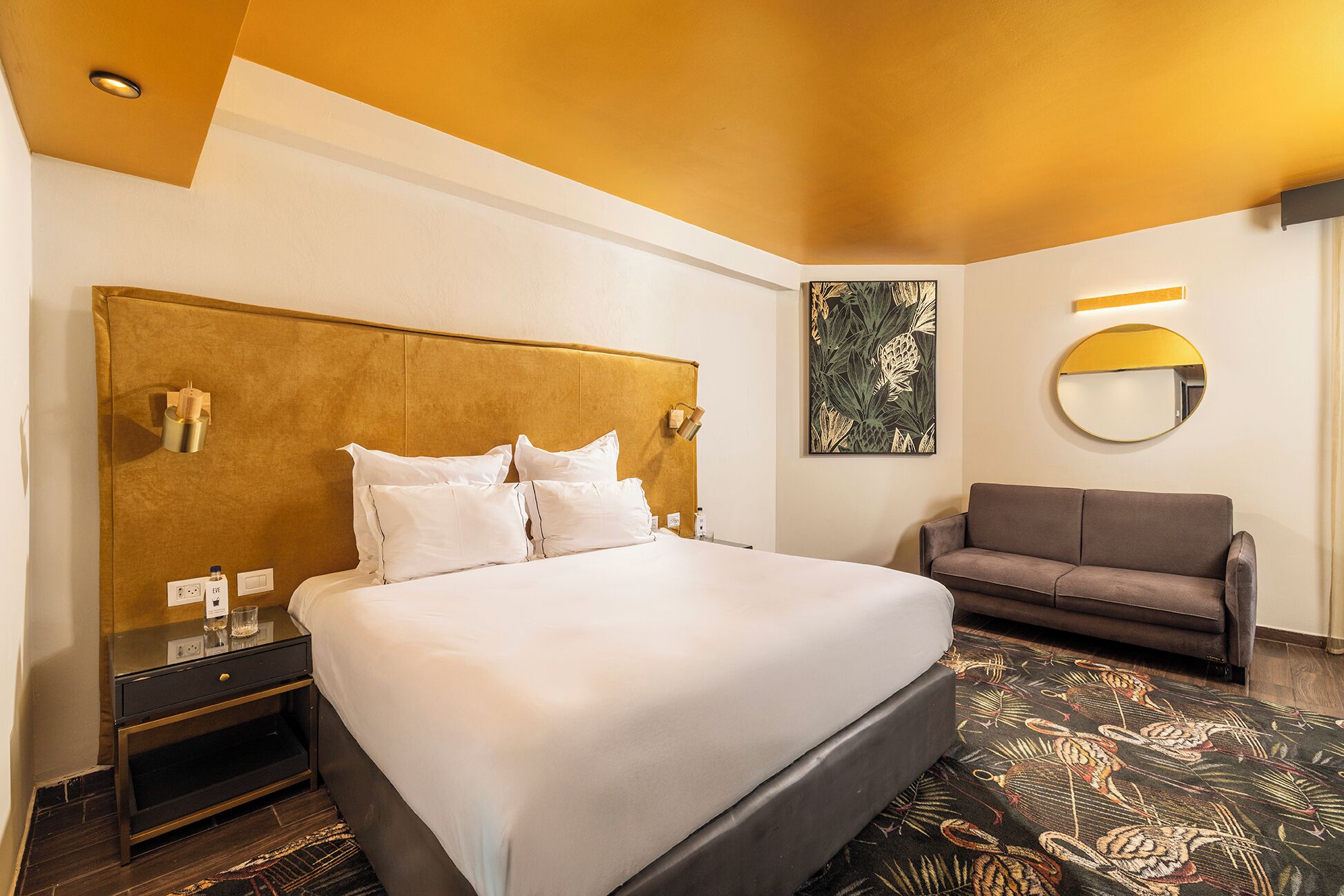 Een hotelkamer met een groot bed, een slaapbank, een ronde spiegel en een kunstwerk aan de muur.