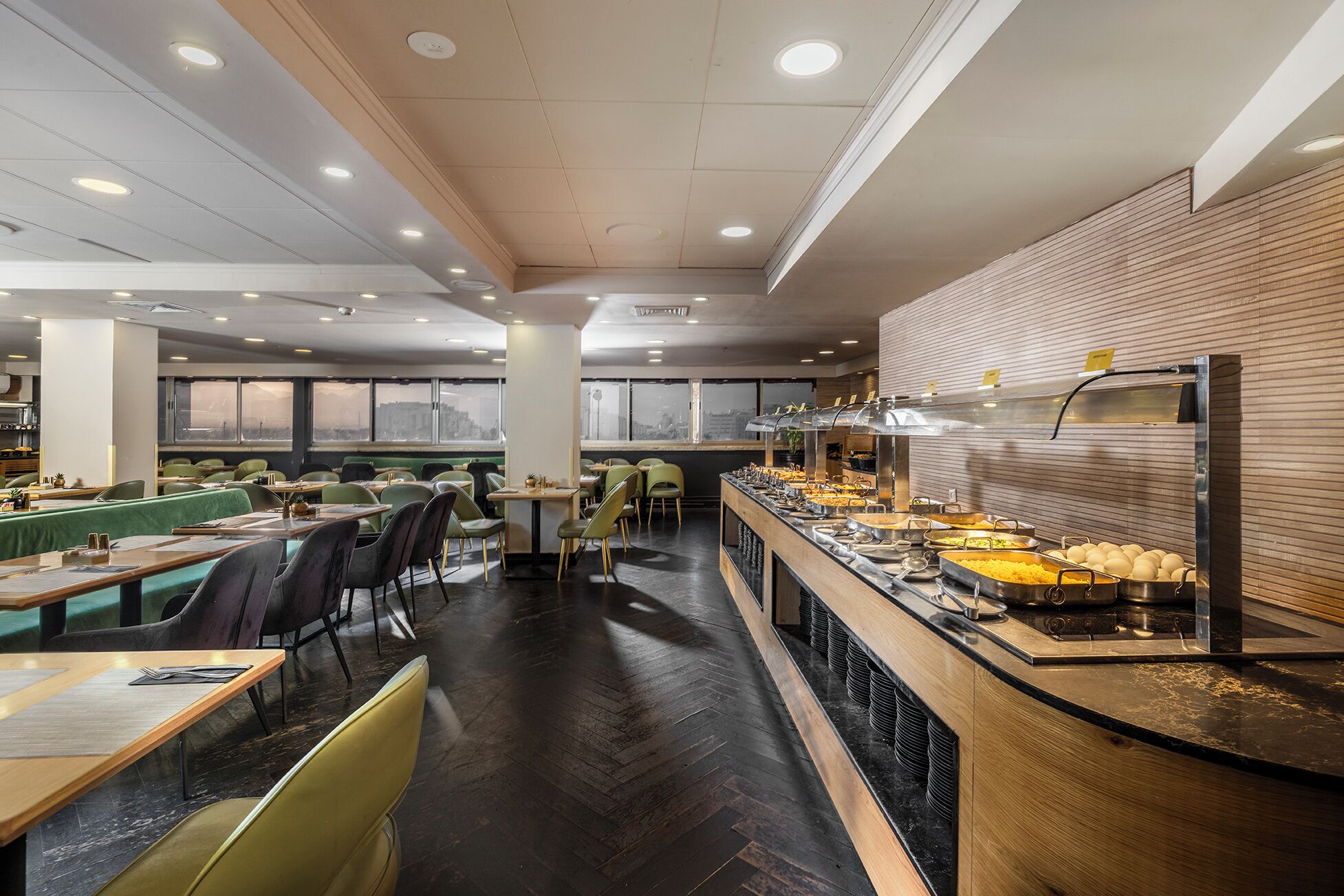 A modern dining area with multiple tables and chairs, a buffet counter with various food items, and large windows providing views of the city.
