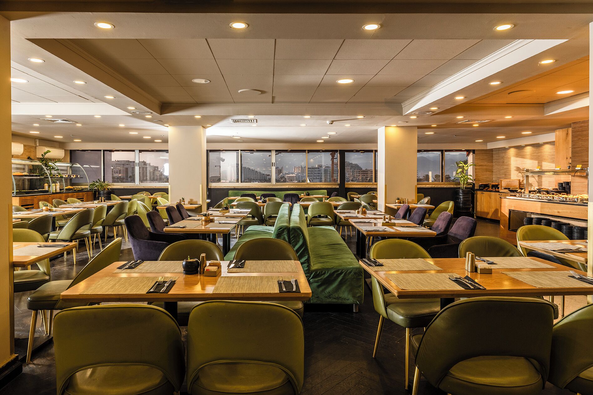 A spacious dining area with multiple tables and chairs, large windows offering a city view, and a kitchen area visible in the background.