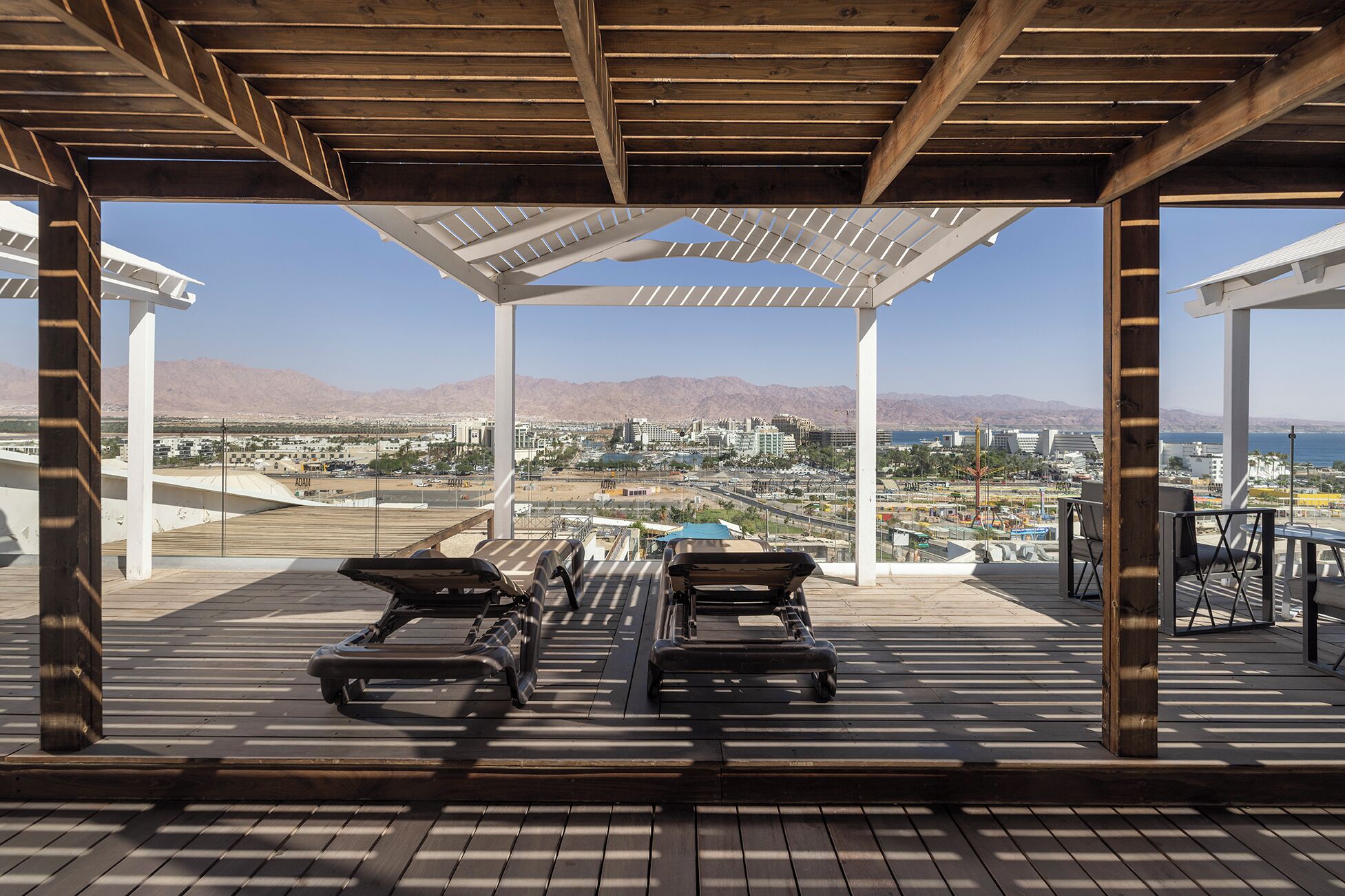 Un espace extérieur abrité comprenant deux chaises longues et une table, offrant une vue sur un paysage urbain avec des montagnes en arrière-plan.