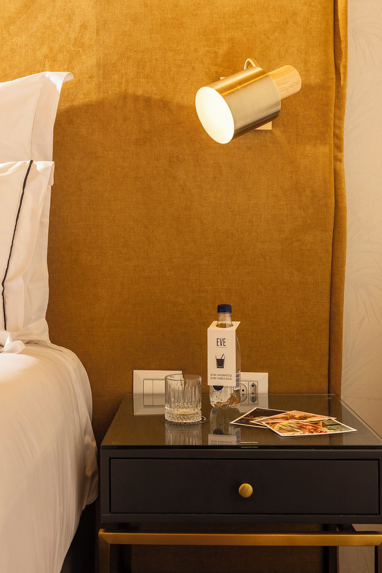 A hotel room with a bed, a nightstand, a bottle of EVE water, a glass, and a menu.