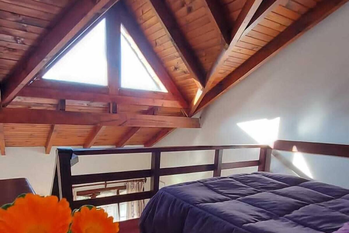 A bedroom with a sloped ceiling, a bed with a purple blanket, and a vase with flowers on a table.