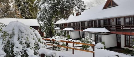 A snow-covered building with a wooden deck, large windows, and a snow-covered tree in the foreground.