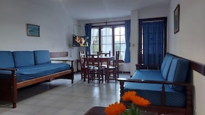 A living room with a sofa, a dining table, and chairs. There is a television mounted on the wall.