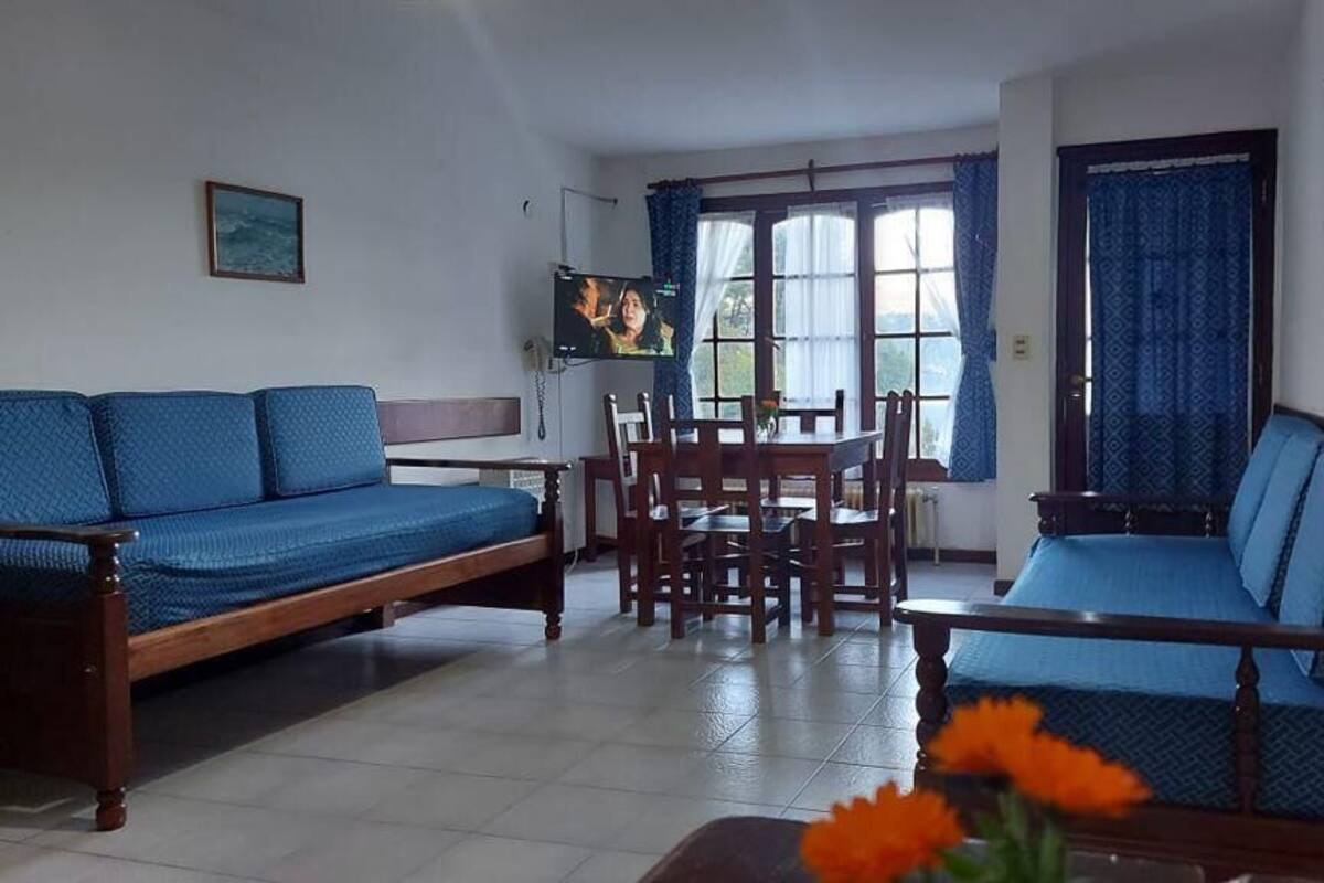 A living room with a sofa, a dining table, and chairs. There is a television mounted on the wall.
