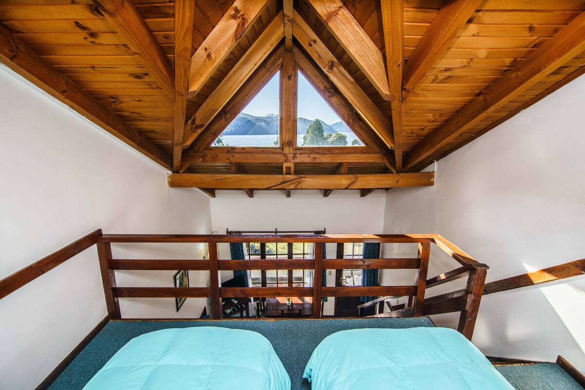 A room with a wooden ceiling and a view of a lake through a triangular window. There are two beds with blue bedding.