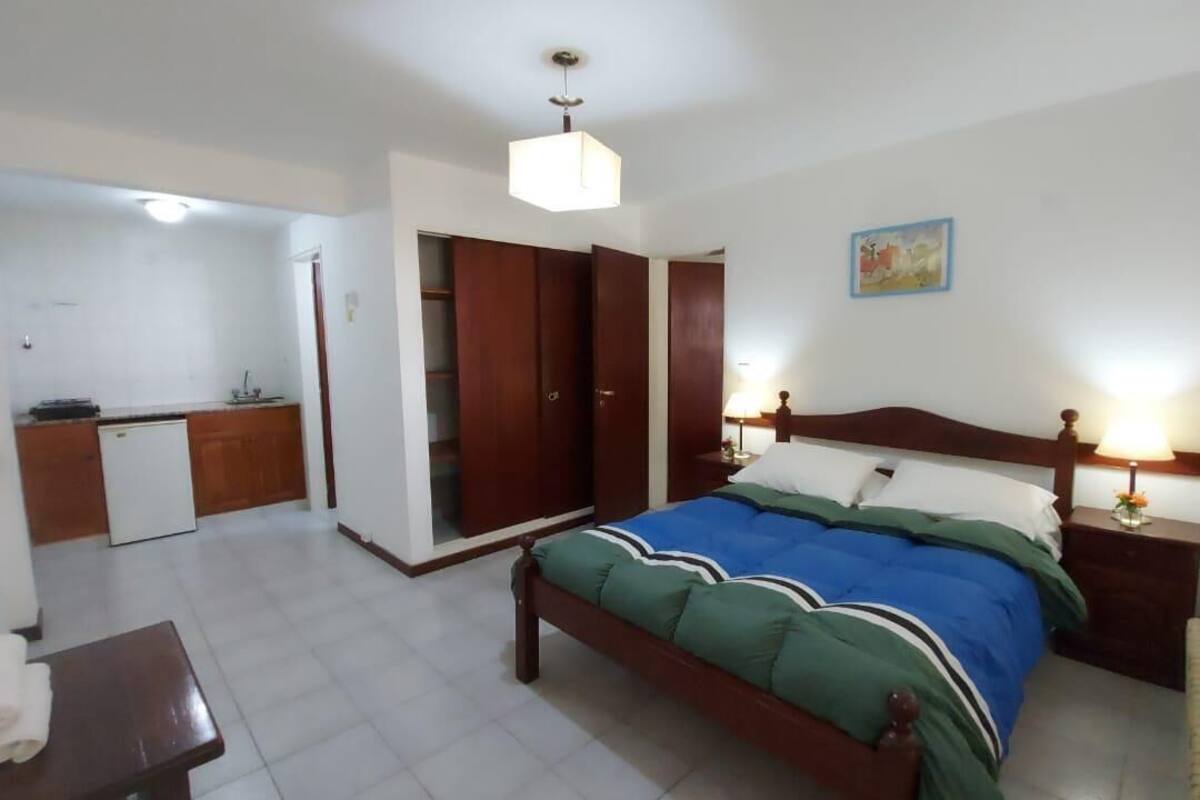 A bedroom with a bed, bedside table, lamp, wardrobe, and a small kitchenette area.
