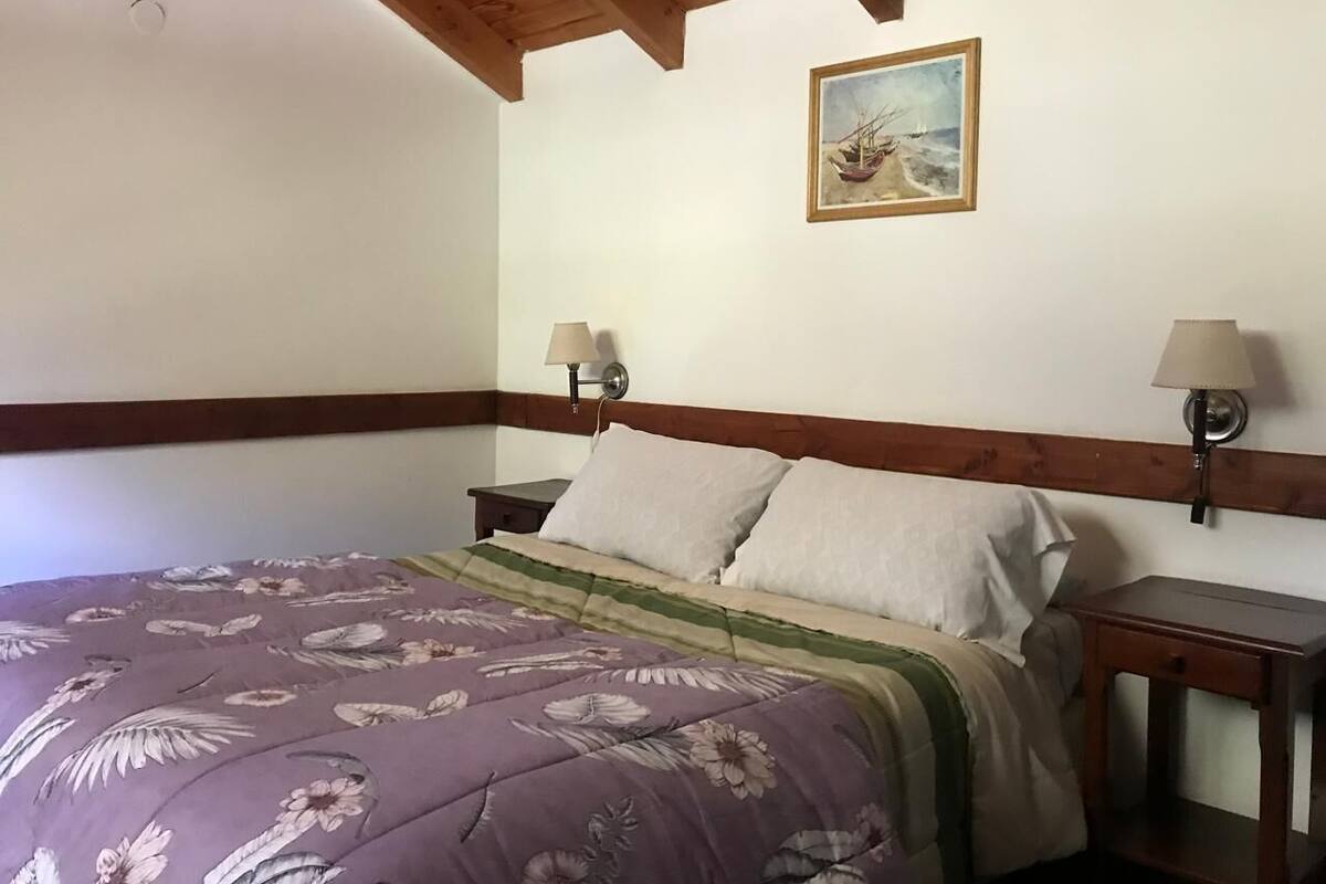 A bedroom with a bed, two bedside tables, a painting on the wall, and a wooden ceiling.