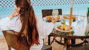 A person seated at an outdoor table with a view of a pool, dining table with plates of food, and a glass of water.