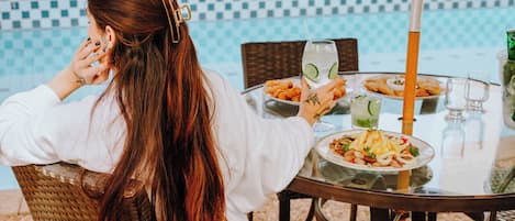 A person seated at an outdoor table with a view of a pool, dining table with plates of food, and a glass of water.