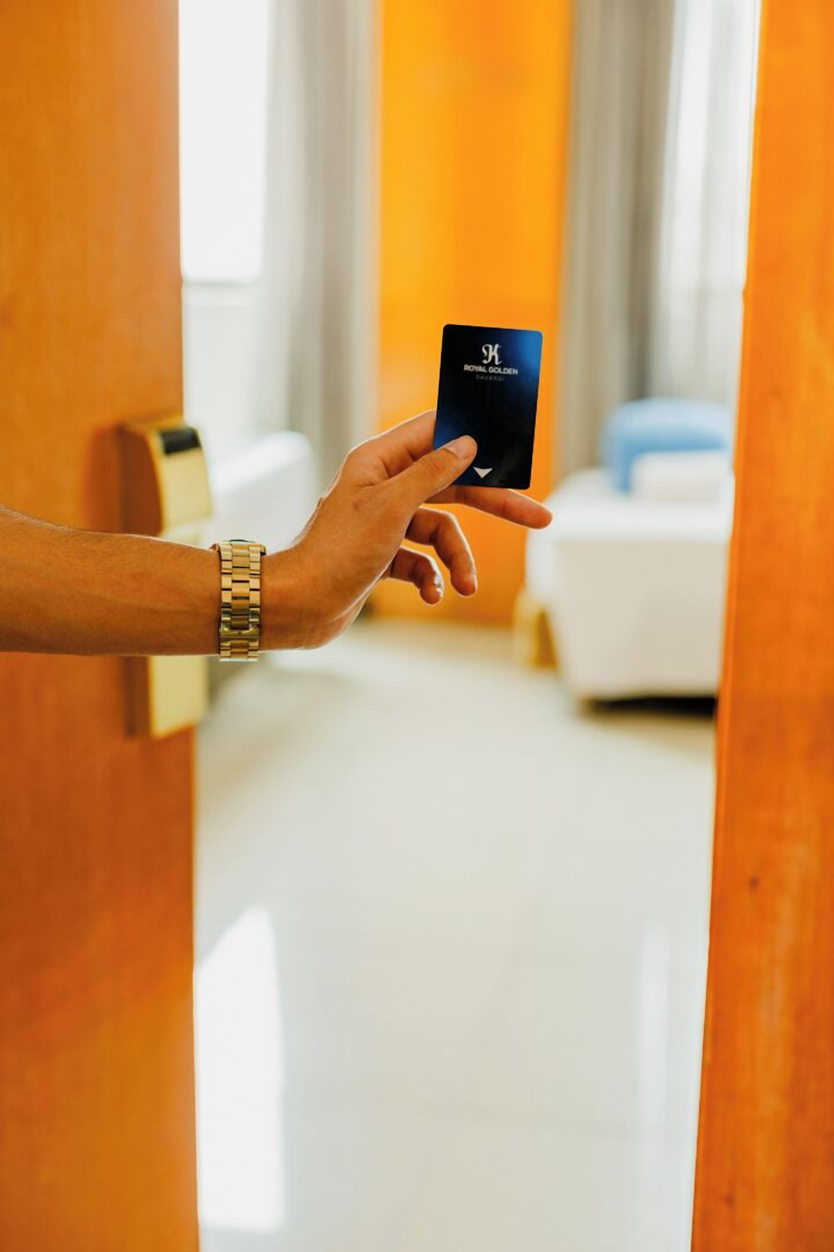 Eine Hand hält eine schwarze Karte mit dem Aufdruck "Royal Golden", während sie einen Raum betritt, der ein weißes Bett und eine Holztür zeigt.