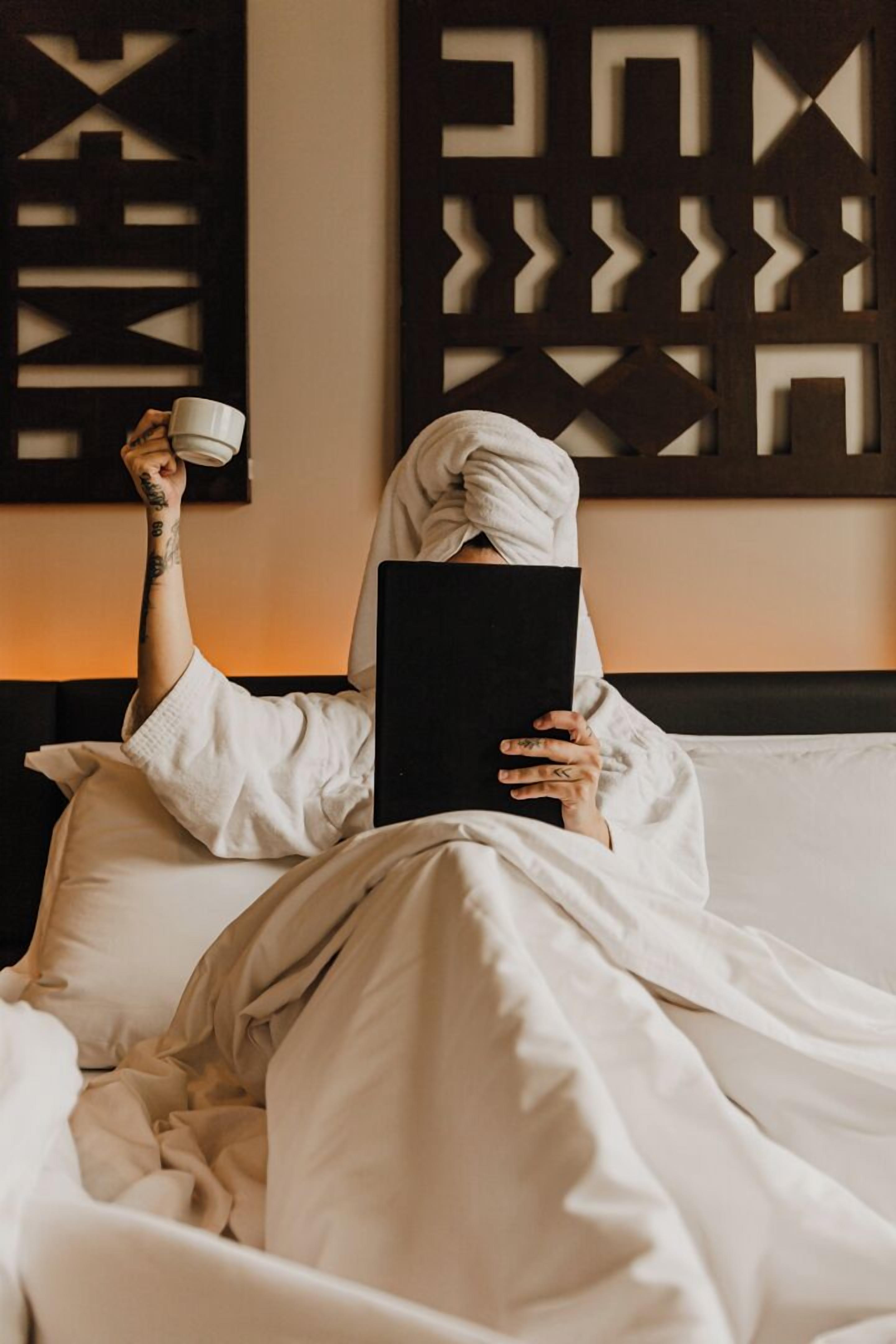 Una persona vestida con una bata blanca sentada en una cama con un libro y una taza, en una habitación con decoración geométrica en las paredes.