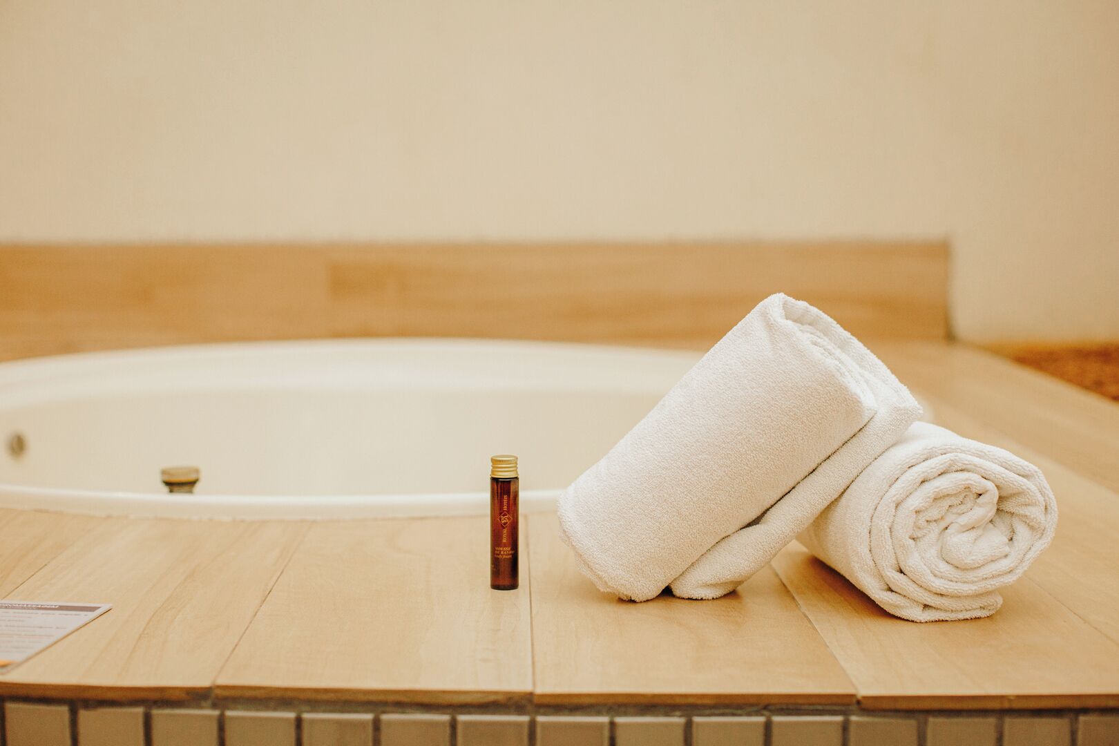 A wooden floor with a white bathtub, a rolled white towel, and a bottle of bath oil.
