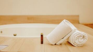 A wooden floor with a white bathtub, a rolled white towel, and a bottle of bath oil.